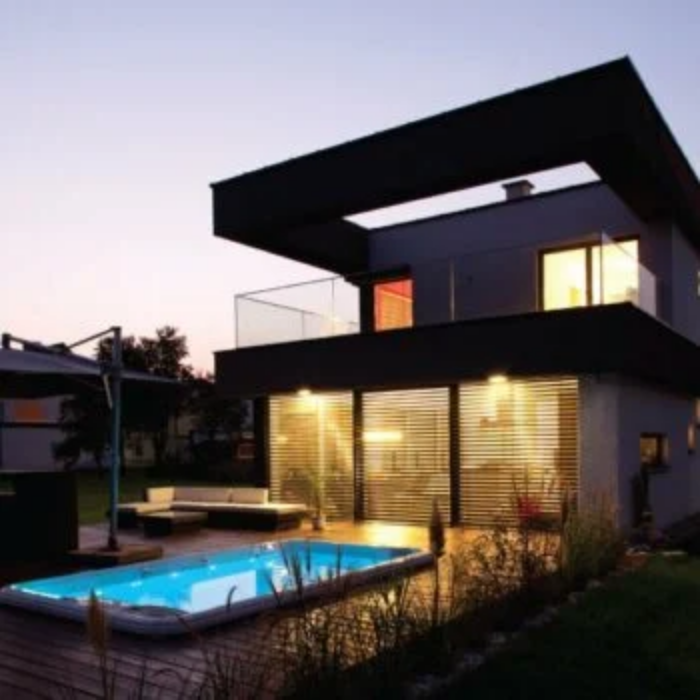 Experience the Hot Tub & Swim Spa Opening at a modern two-story house with large windows, glowing backyard pool, illuminated lights, and a landscaped patio featuring outdoor seating.