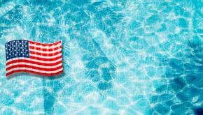 An American flag waves in the upper left corner over a bright blue swimming pool, inspiring Memorial Day pool party ideas with its clear water and visible tile lines below the surface.