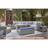 The Naples Beach Grouping 7 piece set features woven gray furniture with gray cushions, a blue-accented bench on a striped rug, surrounded by plants and lanterns, with a large sectional beneath palm trees next to a building.