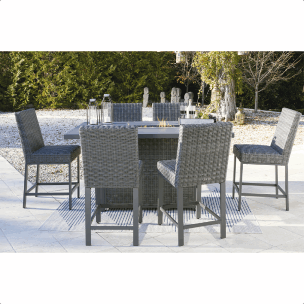 The Palazzo Hill Bar Set 7-piece features six gray wicker chairs and a rectangular fire pit table, arranged on a striped rug atop a tiled patio with lush greenery and stylish garden decor in the background.