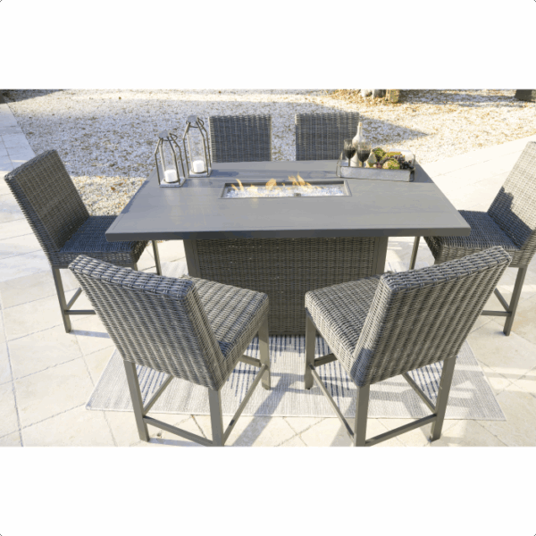The Palazzo Hill Bar Set 7-piece features a modern outdoor bar table surrounded by six wicker chairs. The rectangular table, topped with lanterns, bottles, and fruit, sits on a paved area with gravel visible in the background.