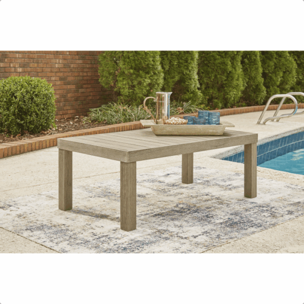The Silo Point Grouping 5 piece wooden outdoor coffee table is set on a patterned rug by the pool, holding a tray with a pitcher and two glasses. Green shrubs and a brick wall form the background.