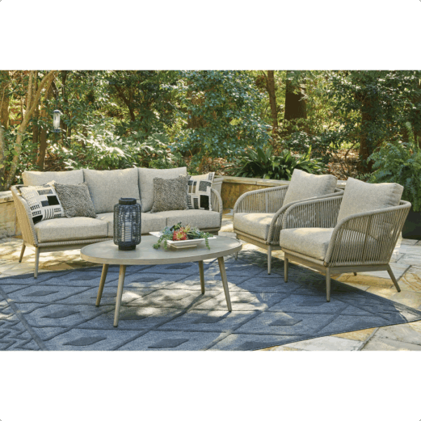 The Swiss Valley Grouping 5 piece features a cushioned sofa, two rope-detailed armchairs, and a round coffee table with accessories, all arranged on a blue rug in a lush garden.