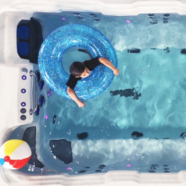 A child in a black shirt floats in the EP14 Swim Spa – Silver Marble Shell/Grey Cabinet on a blue glittery inner tube. A colorful beach ball sits nearby, as clear water shimmers with blue and purple reflections.
