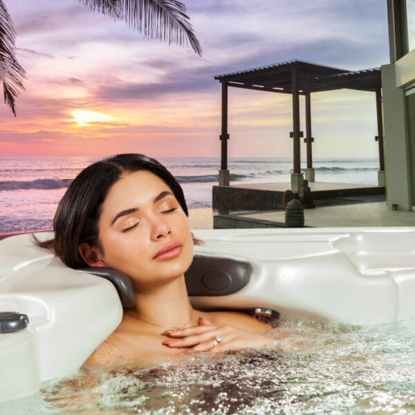 A woman unwinds in a TAHITI Glacier Mountain Shell/Black Cabinet hot tub, eyes closed, hands on her chest, with an ocean sunset, palm fronds, and a modern outdoor pergola as the backdrop.