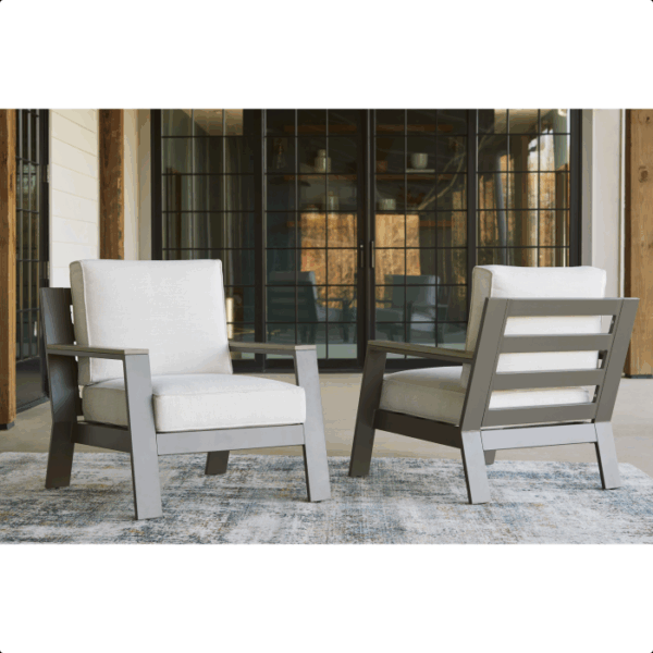 The Tropicava Grouping 4-piece set features two modern lounge chairs with light gray cushions and dark metal frames, arranged on a patterned rug in a spacious room with large glass doors and wooden beams.