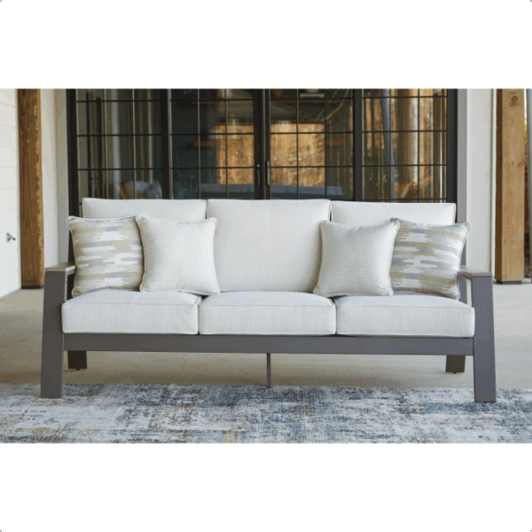 The Tropicava Grouping 4-piece features a modern gray sofa with light cushions and patterned throw pillows, set on a textured rug in front of large windows with black frames.