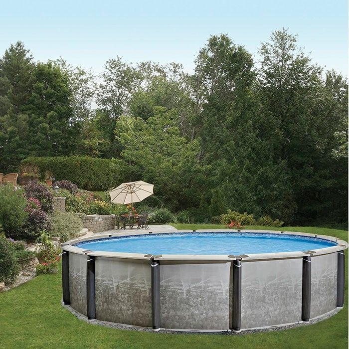 The 24 x 54 Pinnacle Pool Package is set up on a grassy lawn, surrounded by lush trees and landscaping, with a nearby patio area featuring an umbrella and outdoor seating.