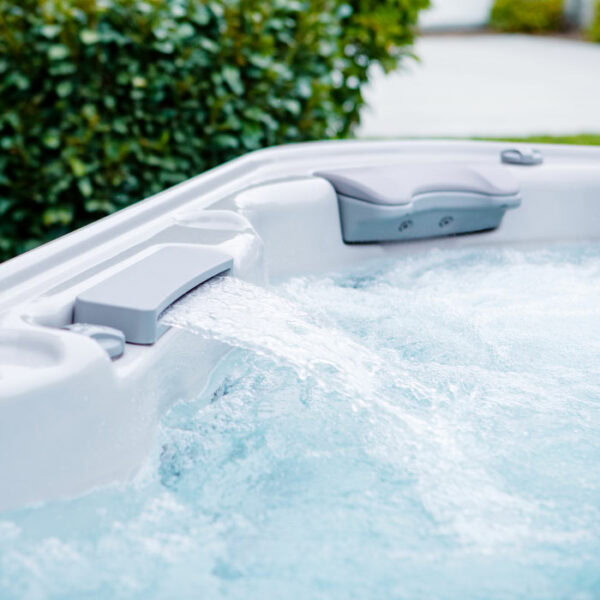 Close-up of water flowing from a spout into the X8 SELECT KIT hot tub (Almond Shell / Acorn Cabinet) outdoors, with green foliage in the background.