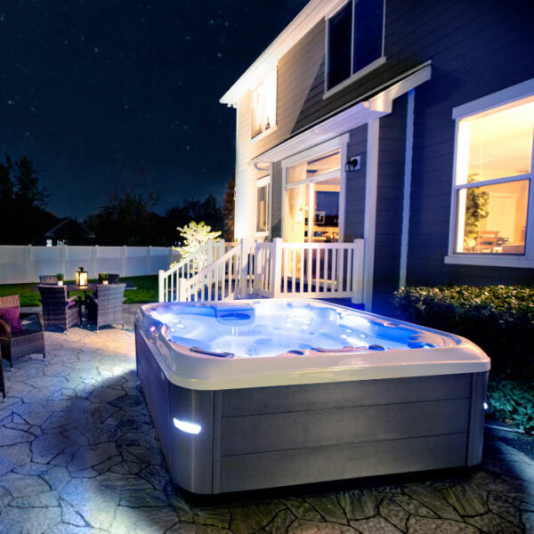 A backyard at night with the X8 SELECT KIT - Almond Shell / Acorn Cabinet hot tub glowing near a patio set. The house has large windows and a porch with steps leading to the well-lit yard.