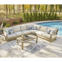 The Silo Point Grouping 5 piece beige outdoor sectional sofa with blue pillows surrounds a wooden coffee table on a rug beside a pool, set against trees and greenery.