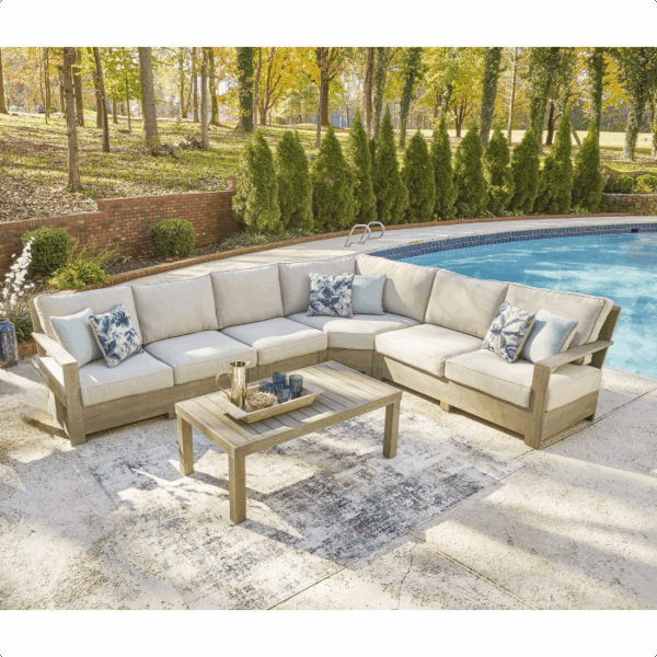 The Silo Point Grouping 5 piece beige outdoor sectional sofa with blue pillows surrounds a wooden coffee table on a rug beside a pool, set against trees and greenery.