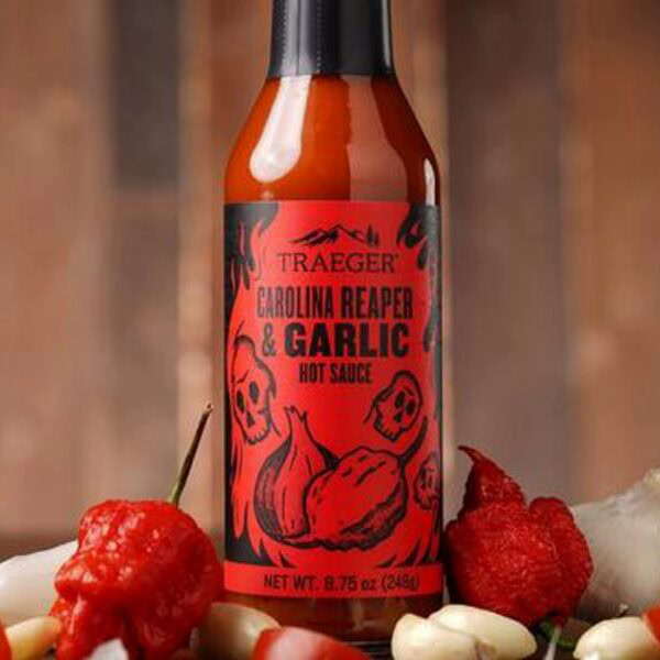 A bottle of Traeger Carolina Reaper & Garlic Hot Sauce sits on wood, surrounded by red chili peppers and garlic. Its red label features black skulls, garlic, and pepper graphics.