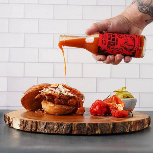 A hand pours Traeger Carolina Reaper & Garlic Hot Sauce onto a fried chicken sandwich with slaw on a wooden board, beside sliced pickles and red peppers, set against a white tile background.
