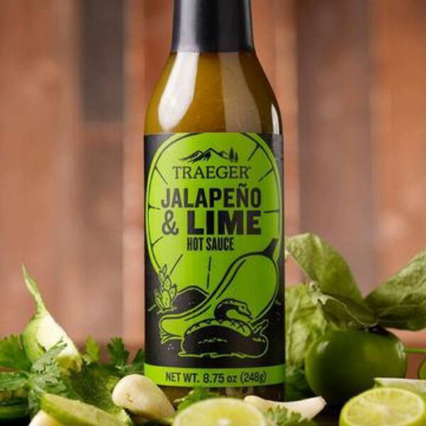 A bottle of Traeger Jalapeño & Lime Hot Sauce sits on a wooden surface, surrounded by fresh lime, jalapeños, cilantro, and garlic with a blurred brown background.