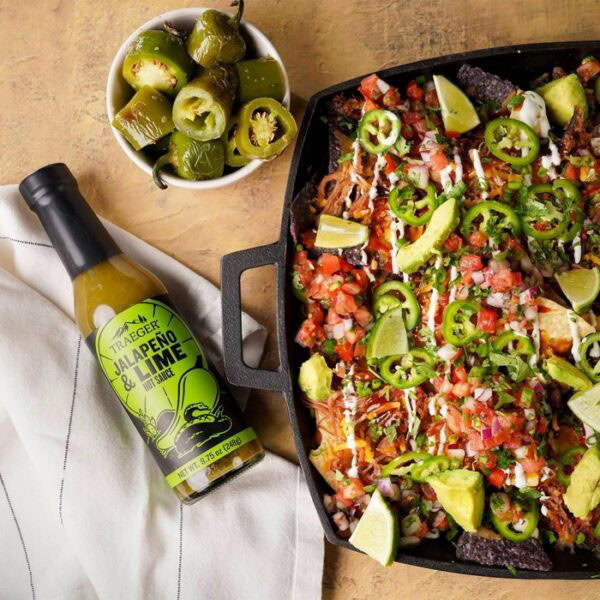 A tray of loaded nachos with jalapeños, avocado, and pico de gallo is served beside a bowl of pickled jalapeños and a bottle of Traeger Jalapeño & Lime Hot Sauce on a tan surface.