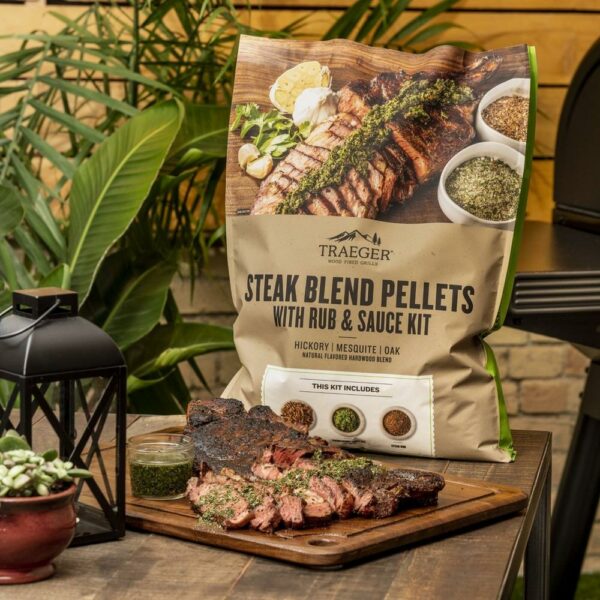 A bag of Traeger Summer Steak Blend Pellets with a rub and sauce kit sits behind a wooden cutting board featuring sliced grilled steak and a bowl of green sauce. Nearby on the table are a lantern and a small potted plant.