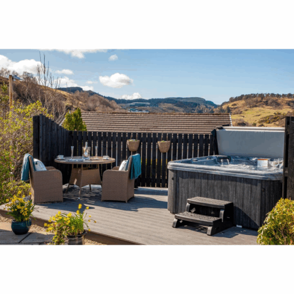 A cozy outdoor deck showcases the Lanai – Midnight Canyon Shell/Black Cabinet hot tub, two wicker chairs with a small drink table, potted plants, and scenic rolling hills beneath a blue sky with clouds.