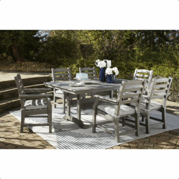 The Visola Dining Set 7 piece, featuring a gray rectangular table and six chairs, is placed on a striped rug on a stone patio. The table is adorned with white flower vases, plates, and cake, with greenery and stone steps in the background.